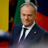 Donald Tusk, w garniturze i krawacie, z przypinką w kształcie serca, stoi na tle flag. Jego rola w Unii Europejskiej jest kluczowa.