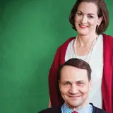 Uśmiechnięta kobieta w perłach i czerwonym szalu stoi za mężczyzną w pasiastym krawacie.