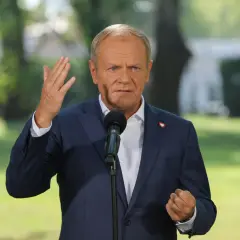 Donald Tusk w garniturze, przemawiający do mikrofonu, z serduszkiem na klapie.