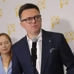 Szymon Hołownia w okularach i garniturze przemawia przy mikrofonie, za nim kobieta i logo Polska 2050.