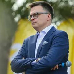 Szymon Hołownia w garniturze i okularach, z mikrofonem w ręku, na tle żółtych flag.