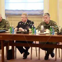Czterech mężczyzn w mundurach siedzi przy drewnianym stole, przed nimi butelki wody i filiżanki.