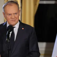 Donald Tusk w garniturze przemawia przy mikrofonie, obok polska flaga.