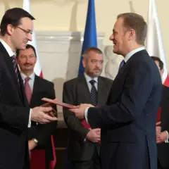 Mateusz Morawiecki przekazuje dokument Donaldowi Tuskowi, stojąc przed polskimi i unijnymi flagami.