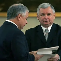 Dwóch mężczyzn w garniturach, jeden podaje drugiemu dokumenty.