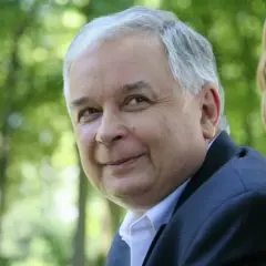 Lech Kaczyński w garniturze siedzi na tle zieleni i wiklinowego fotela.