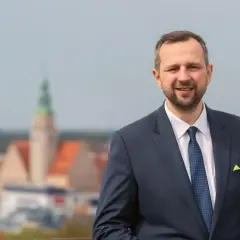 Mężczyzna w garniturze uśmiecha się na tle panoramy miasta z wieżą.