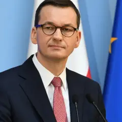 Mateusz Morawiecki w garniturze i okularach, na tle flag Polski i UE.