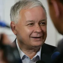 Lech Kaczyński podczas wywiadu, w tle mikrofony i niewyraźne postacie.