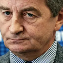 Mężczyzna w garniturze, w tle niebieska krata z białymi literami. Czy to on wyjaśnia, dlaczego posłowie PiS zostali oskarżeni o nadużywanie lotów rządowych?