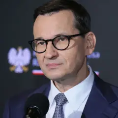 Mateusz Morawiecki w okularach i garniturze przed mikrofonem, na tle godła Polski.
