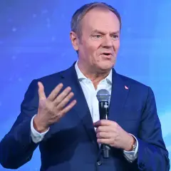 Donald Tusk w garniturze, przemawiający do mikrofonu na tle niebieskiego ekranu.