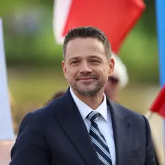 Rafał Trzaskowski w garniturze i krawacie, uśmiecha się na tle flag.