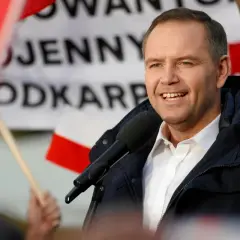 Mężczyzna w białej koszuli i granatowej kurtce przemawia do mikrofonu na tle flag.