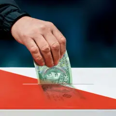 Dłoń wrzuca banknot 100 zł do urny w barwach narodowych.