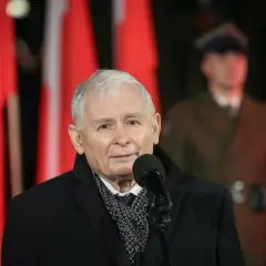 Jarosław Kaczyński przemawia przy mikrofonie na tle polskich flag i żołnierza.