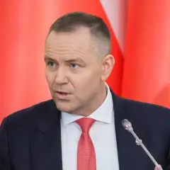 Mężczyzna w garniturze i czerwonym krawacie przemawia na tle czerwonych flag. Dyskutuje, dlaczego prezydent zapowiada veto wobec ustawy o wyborze sędziów do KRS.