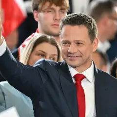 Rafał Trzaskowski w garniturze i czerwonym krawacie, pozdrawia tłum, unosząc rękę ze znakiem zwycięstwa.