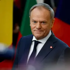 Donald Tusk, w garniturze i krawacie, z przypinką w kształcie serca, stoi na tle flag. Jego rola w Unii Europejskiej jest kluczowa.