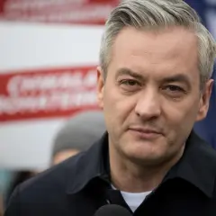 Robert Biedroń, polityk, w czarnej kurtce, z siwymi włosami. W tle czerwone napisy.