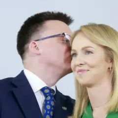 Szymon Hołownia szepcze coś do ucha Urszuli Brzezińskiej Hołowni. W tle godło Polski.