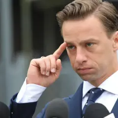 Krzysztof Bosak w garniturze, z palcem przy skroni, przed mikrofonami.