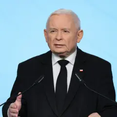 Jarosław Kaczyński w czarnym garniturze przemawia przy mikrofonie na tle niebieskiego tła.