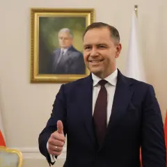 Mężczyzna w garniturze pokazuje kciuk w górę na tle portretu i flag Polski.