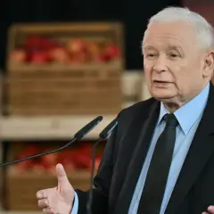 Jarosław Kaczyński w garniturze przemawia przy mikrofonach, w tle skrzynki z jabłkami.