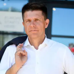 Ryszard Petru w białej koszuli, z marynarką na ramieniu, na tle budynku.