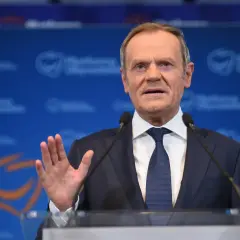 Donald Tusk w garniturze i krawacie przemawia przy mównicy z mikrofonami.