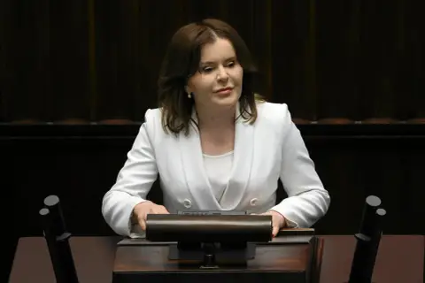 Anna Krupka w białej marynarce przy mównicy. Czy wiceprezes PiS nie dostaje pieniędzy za funkcję?