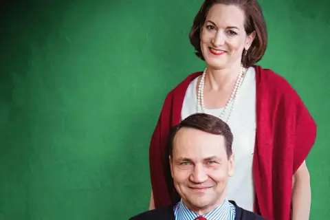 Uśmiechnięta kobieta w perłach i czerwonym szalu stoi za mężczyzną w pasiastym krawacie.