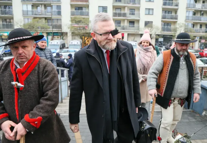 Grzegorz Braun w czarnym płaszczu i z opaską na oku, w otoczeniu mężczyzn w tradycyjnych strojach.