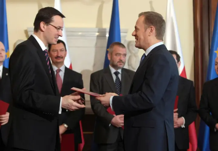 Mateusz Morawiecki przekazuje dokument Donaldowi Tuskowi, stojąc przed polskimi i unijnymi flagami.