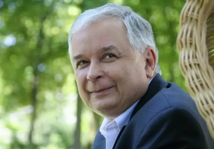 Lech Kaczyński w garniturze siedzi na tle zieleni i wiklinowego fotela.
