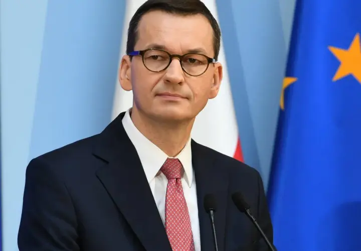 Mateusz Morawiecki w garniturze i okularach, na tle flag Polski i UE.