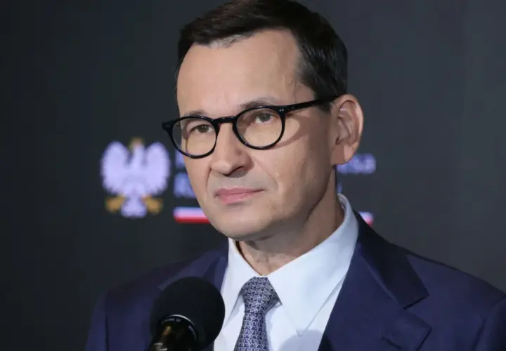 Mateusz Morawiecki w okularach i garniturze przed mikrofonem, na tle godła Polski.