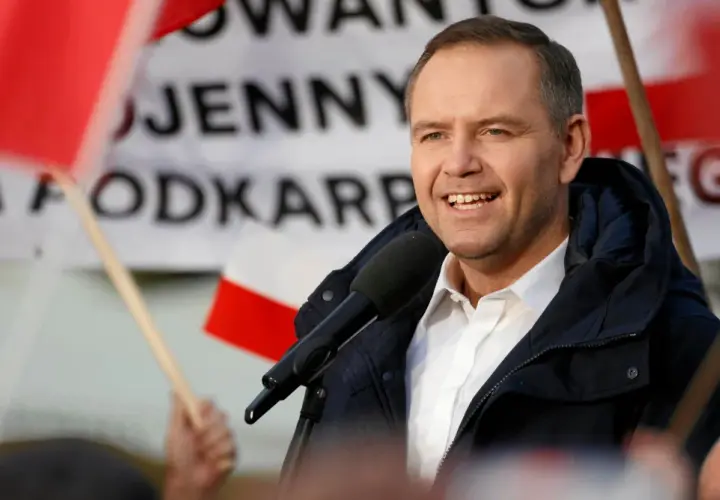 Mężczyzna w białej koszuli i granatowej kurtce przemawia do mikrofonu na tle flag.