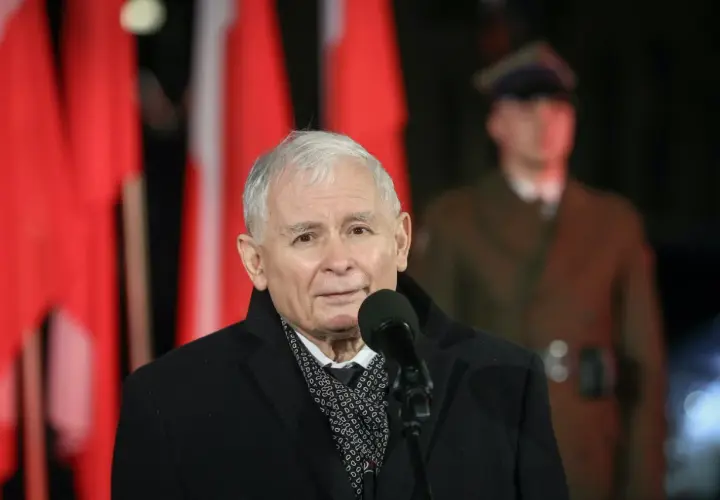 Jarosław Kaczyński przemawia przy mikrofonie na tle polskich flag i żołnierza.