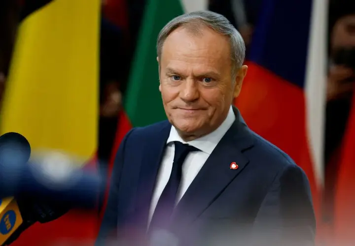Donald Tusk, w garniturze i krawacie, z przypinką w kształcie serca, stoi na tle flag. Jego rola w Unii Europejskiej jest kluczowa.