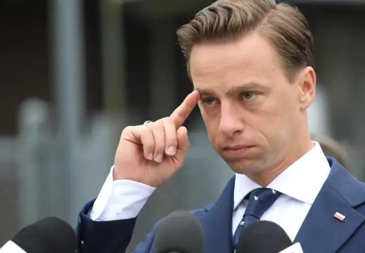Krzysztof Bosak w garniturze, z palcem przy skroni, przed mikrofonami.
