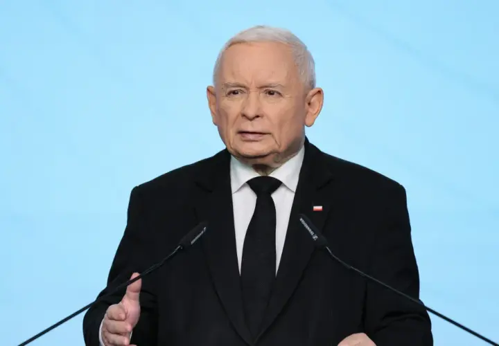 Jarosław Kaczyński w czarnym garniturze przemawia przy mikrofonie na tle niebieskiego tła.