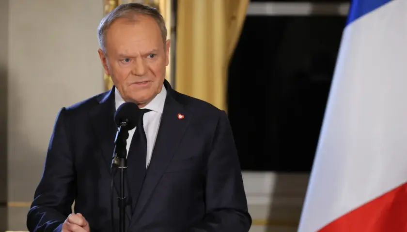 Donald Tusk w garniturze przemawia przy mikrofonie, obok polska flaga.