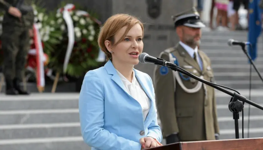Anna Krupka w błękitnej marynarce przemawia przy mikrofonie. W tle żołnierze i wieńce. Ile milionów złotych oszczędności wykazała w oświadczeniu?