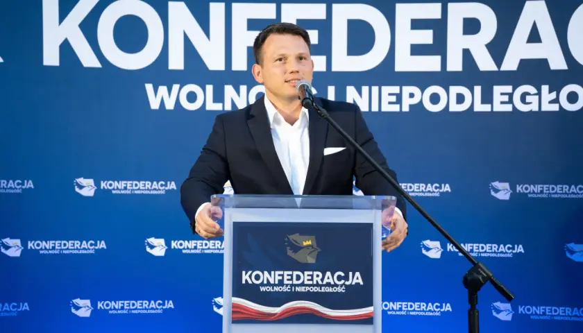 Mężczyzna w garniturze przemawia na tle logo Konfederacji. Czy to zapowiedź sekretów negocjacji Konfederacji z rolnikami w sprawie blokad granic w marcu 2026?