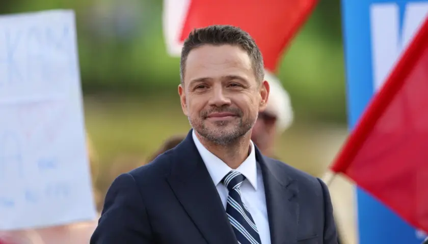 Rafał Trzaskowski w garniturze i krawacie, uśmiecha się na tle flag.