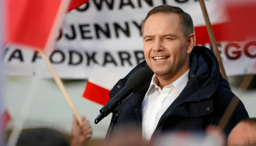 Mężczyzna w białej koszuli i granatowej kurtce przemawia do mikrofonu na tle flag.