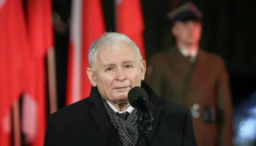 Jarosław Kaczyński przemawia przy mikrofonie na tle polskich flag i żołnierza.