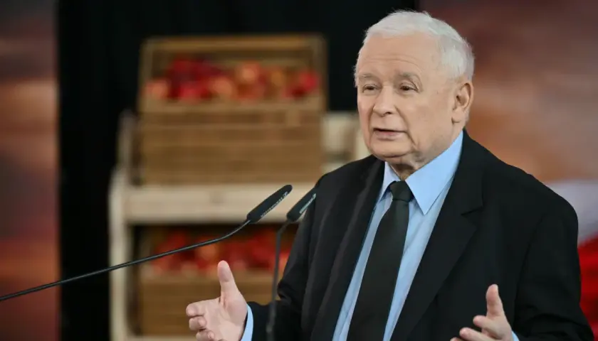 Jarosław Kaczyński w garniturze przemawia przy mikrofonach, w tle skrzynki z jabłkami.